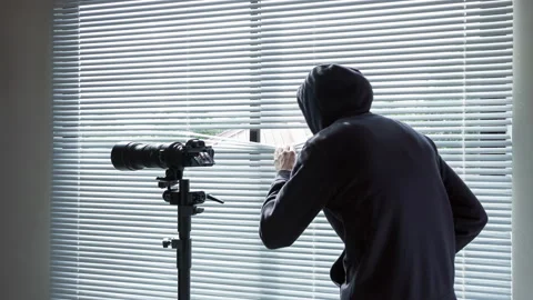The man in black stands against the window and looks through the blinds. 库存影片 97701301
