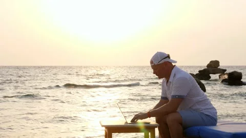 Man in blazer hat, typing using laptop, sitting on the beach by the sea, at Stock Footage 136756326