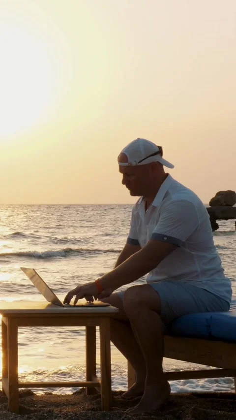 Man in blazer hat, typing using laptop, sitting on the beach by the sea, at Stock Footage 313984360