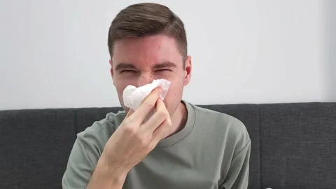Man blowing nose with tissue while sitting on a sofa. Illness and allergy Stock Footage 282609572