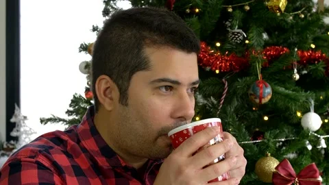 Man blowing red mug and drinking hot beverage in front of Christmas tree Stock Footage 140675502