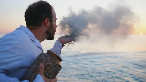 A man blows ashes from the palm of his hand with ashes from a cremation urn slow Stock Footage 247175129