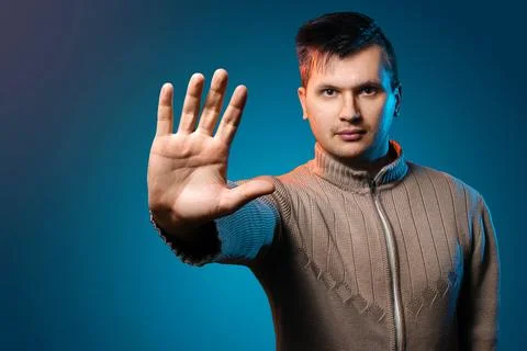 A man on a blue background in a yellow jacket shows his hand to stop. Serious Foto stock