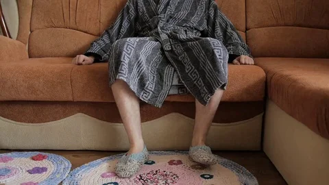 The man in blue bathrobe sitting on brown sofa. Knitted slippers on his feet Stock Footage 69218506