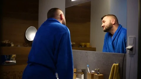 Man in blue bathrobe standing in front of the mirror and having fun with himself Stock Footage 73789009