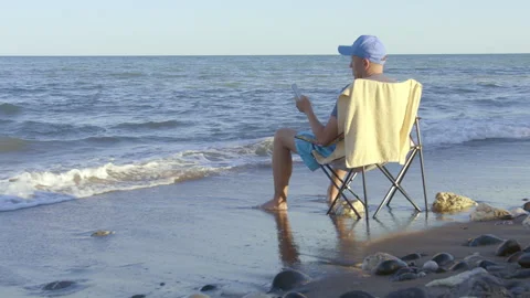 A man in a blue cap sits on the beach on a beach chair with a cell phone Stock Footage 267761116