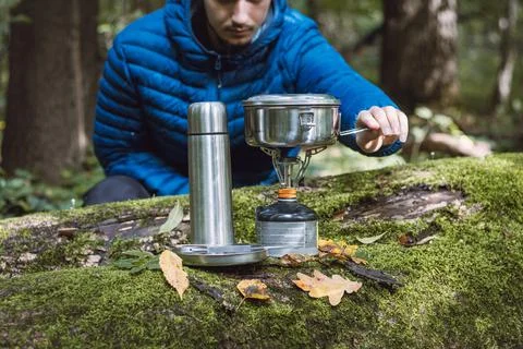 Man in a blue down jacket boils water for tea and food on a portable stove an Stock Photos