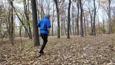 Man in blue jacket and gray beanie running along a forest path covered with Stock Footage 316843517
