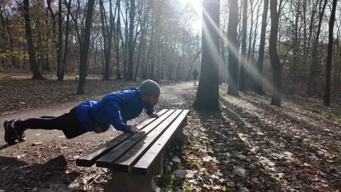 Man in a blue jacket and grey hat does push-ups against a bench in the forest on Stock Footage 316834825