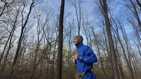 Man in a blue jacket and grey hat jogs through the forest in autumn, training Stock Footage 316835834