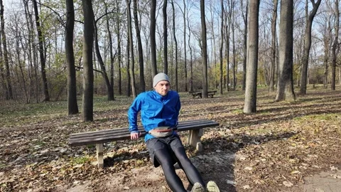 Man in a blue jacket and grey hat does tricep dips on a bench in the forest on a Stock Footage 316836208
