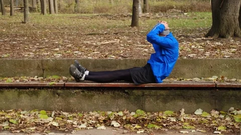 Man in blue jacket doing sit-ups on bench in autumn park, exercising abs during Stock Footage 315584703