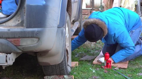 A man in a blue jacket installs a jack under a car in the countryside. Stock Footage 143575116
