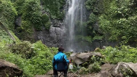 Man in Blue Jacket Watching Waterfall in Lush Green Forest, Wide Static Shot Video stock 324851338