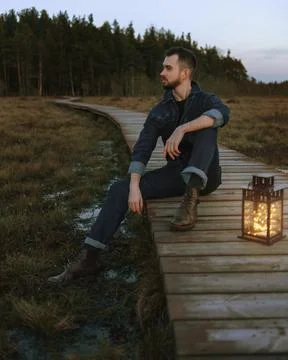 A man in a blue overalls and brown boots sits on a wooden one in the forest. Stock Photos