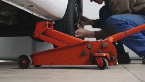 A man in blue overalls is changing a tire on a lifted car indoors. Stock Footage 182916409
