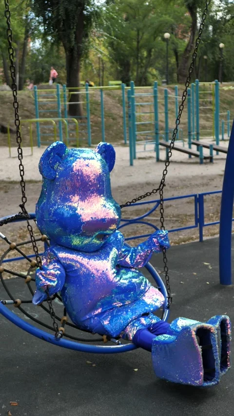 Man in blue, shiny mascot bear swinging on a swing in a city park. Carnival Stock Footage 289674191