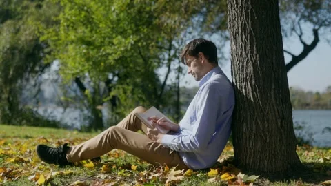 Man in blue shirt sitting under tree reading book in park, cinematic Stock Footage 332298776