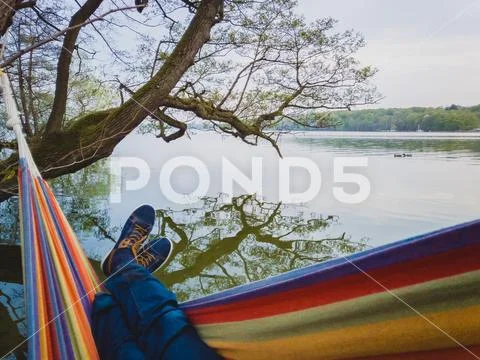 Man with blue shoes and jeans swings in colorful hammock hangs on shore of the Stock Photos