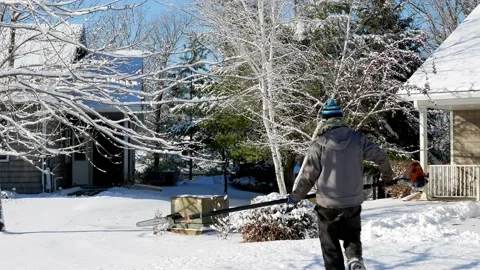 Man in blue stocking cap walks on snow covered yard carrying a chainsaw Stock Footage 130880304