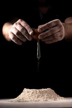 A man breaks an egg into flour for making dough on a table against a dark bac Stock Photos
