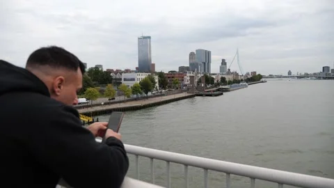 Man on bridge checking phone with city skyline and river in background Vidéo 319910590