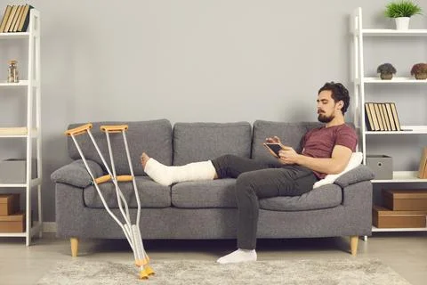Man with a broken leg lying on a sofa and browsing social networks on a digital Fotos Stock