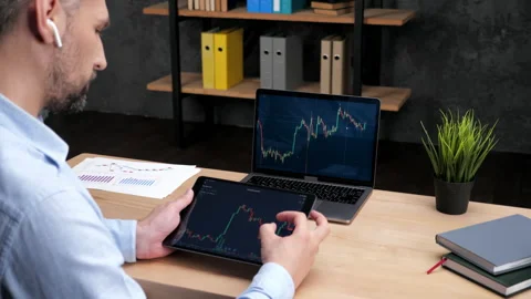 Man broker scrolling tablet with stock exchange chart, laptop with stock market Stock Footage 167018712