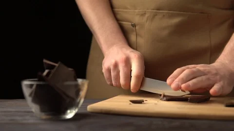 A man in a brown apron with a knife chopping dark chocolate. slow motion Stock Footage 100407430