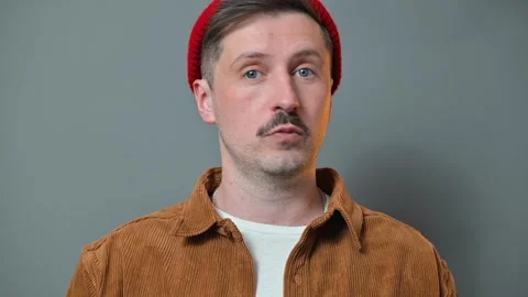 A man in a brown jacket and red beanie makes expressive hand gestures against a 库存影片 337642763