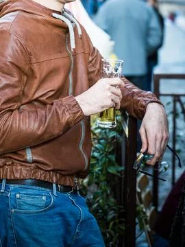 Man in brown leather jacket drinks beer outdoors. Guy holds glass in on hand and Stock Photos
