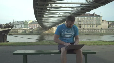 Man is browsing laptop, sitting on bench against the river, steadicam Vídeos de archivo 65205251