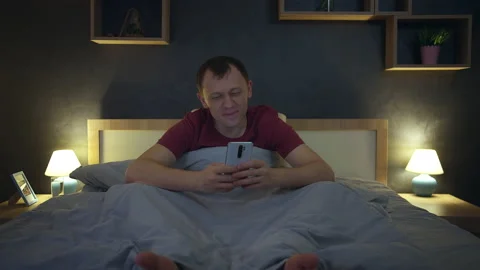 Man browsing mobile apps at night sitting in bed, evening lighting, front view Stock Footage 220680055