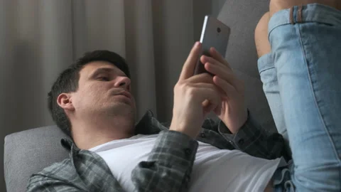 Man is browsing smartphone finding a woman on dating site online lying on sofa. Stock Footage 152812342