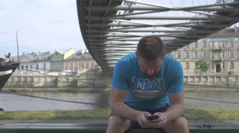 Man is browsing smartphone, sitting on bench against the river. 库存影片 65188497