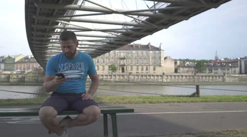 Man is browsing smartphone, sitting on bench against the river, pan shot. 库存影片 65205062