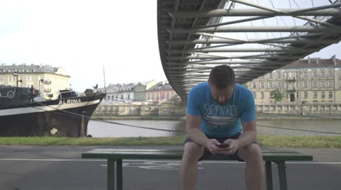 Man is browsing smartphone, sitting on bench against the river, pan shot. Vidéo 65205076