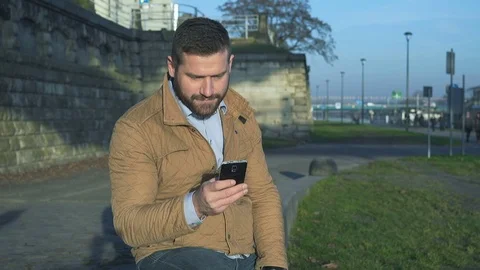 Man browsing smartphone, smiling at camera sitting on wall at boulevar Vidéo 72775255