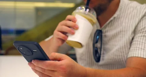 Man browsing smartphone while holding iced drink in cafe, seated at table with 스톡 동영상 344734761