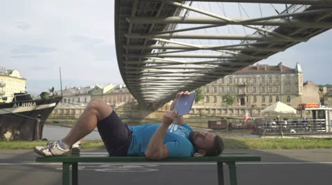 Man is browsing tablet, lying on the bench against the river. Vidéo 65188412