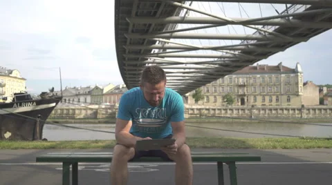 Man is browsing tablet, sitting on bench against the river. 動画素材 65188302