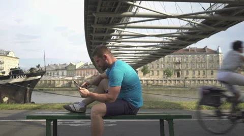 Man is browsing tablet, sitting on bench against the river. 库存影片 65188377