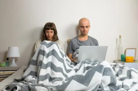 Man browsing web in laptop and angry wife together in bed. Unhappy, dissatisf Stock Photos