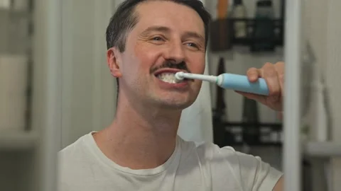 A man brushes his teeth with an electric toothbrush while standing in front of a 库存影片 337111700