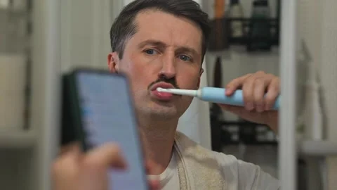 A man brushes his teeth with an electric toothbrush while looking at his 库存影片 337111707