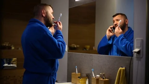 Man brushes his teeth while chatting on cellphone in the bathroom Stock Footage 73789117