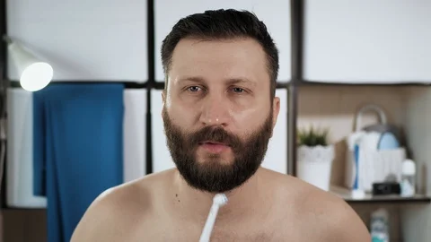 Man is brushing his teeth. Young bearded caucasian guy looks in mirror in Vídeos de archivo 128231817