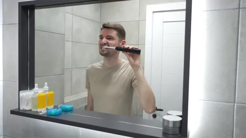Man brushing teeth with electric toothbrush in front of bathroom mirror Video stock 283931512