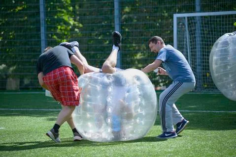 Man in a bubble inside felling upside down on a football field, two men helpi Stock Photos