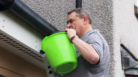 Man with bucket on ladder cleans gutter on tiled roof low angle medium shot Stock Footage 271694625
